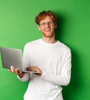 Cheerful redhead freelancer in glasses smiling at camera, working on laptop while standing over green background